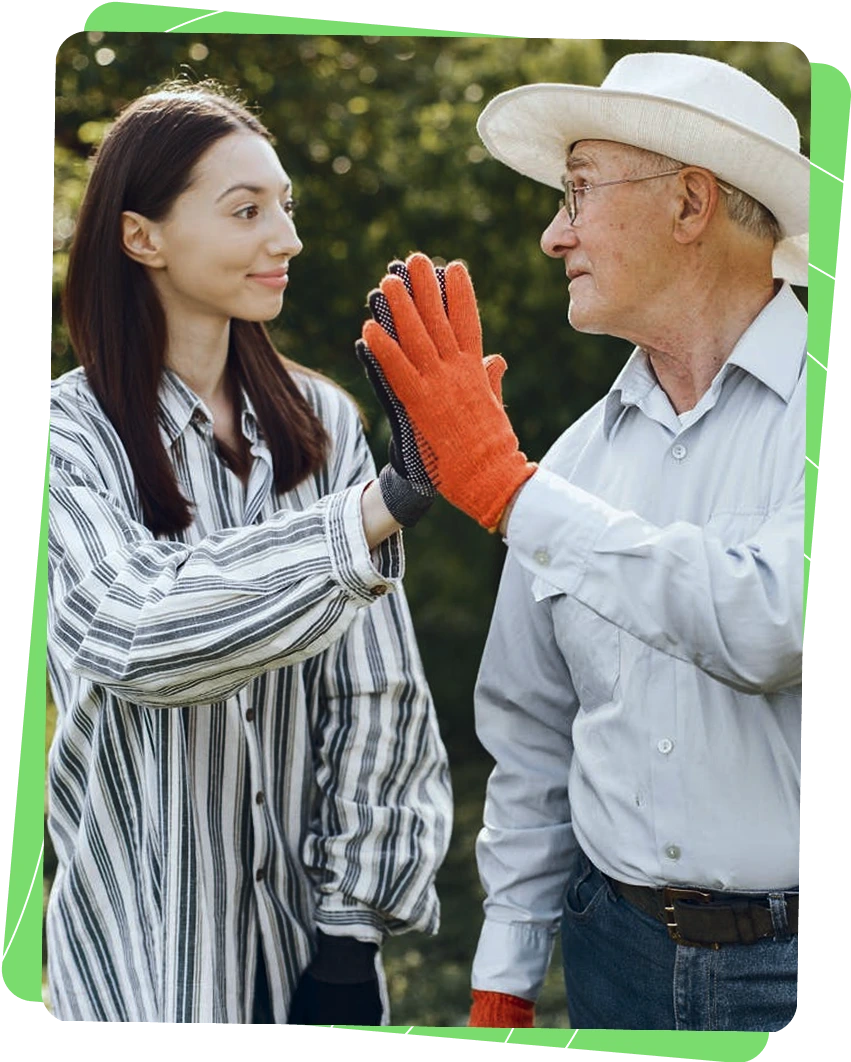 Une jeune volontaire en Service Civique Écologique et un tuteur se félicitent après une action environnementale, portant des gants de jardinage orange. Ils échangent un geste complice dans un cadre naturel verdoyant, illustrant la collaboration intergénérationnelle pour la protection de l'environnement.
